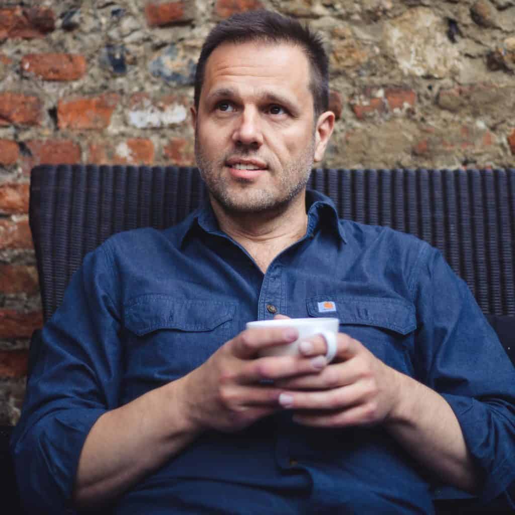 Professional portrait of Adam de-Ste-Croix smiling and holding a coffee cup